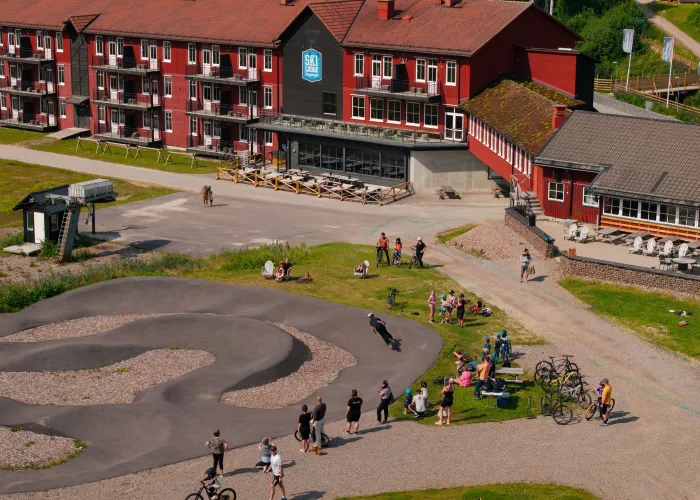 Drönarbild över Ski Lodge på sommaren. Framför Ski Lodge cyklar gäster på en asfalterad cykelbana. Det är en solig dag med gröna områden. 