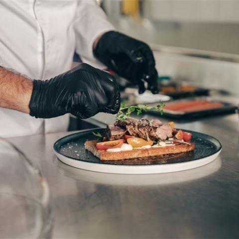 Kock med svart handskar lägger upp mat på tallrik i köket